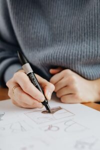 Crop anonymous female artist in casual wear with ring on finger using marker while drawing on paper with drafts at wooden table at work